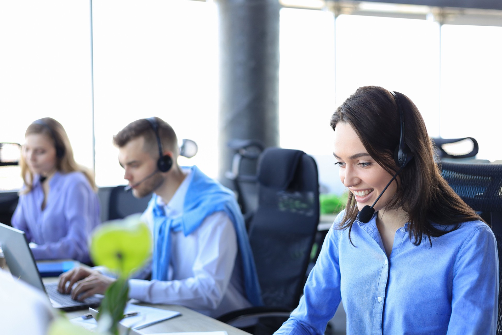 Opératrice de support client féminin avec casque et sourire, avec des collègues en arrière-plan.